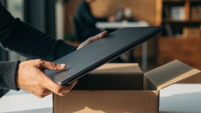 Hands lifting a brand-new graphite black laptop out of its plain cardboard box on a white desk