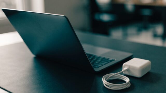 A graphite black laptop on a dark desk mat next to a compact white power adapter with its cable neatly coiled