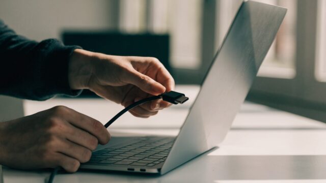 Hands unplugging a charging cable from a slim silver laptop on a white desk