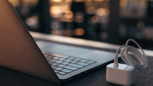 A matte grey laptop on a dark desk mat next to a compact white power adapter with its cable neatly coiled