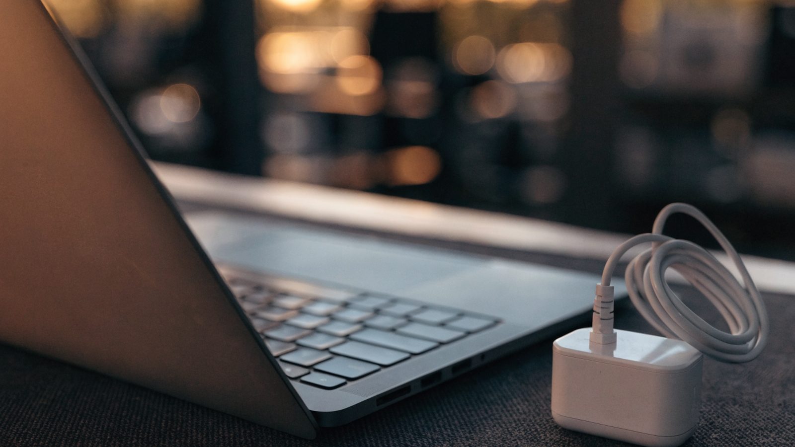 A matte grey laptop on a dark desk mat next to a compact white power adapter with its cable neatly coiled