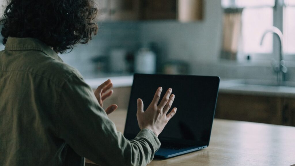 A person seen from behind with hands raised at a dark blue laptop with a dark blank screen on a kitchen table