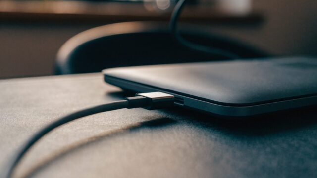 Close-up of a USB-C charger cable plugged into the side port of a graphite black laptop on a dark desk mat