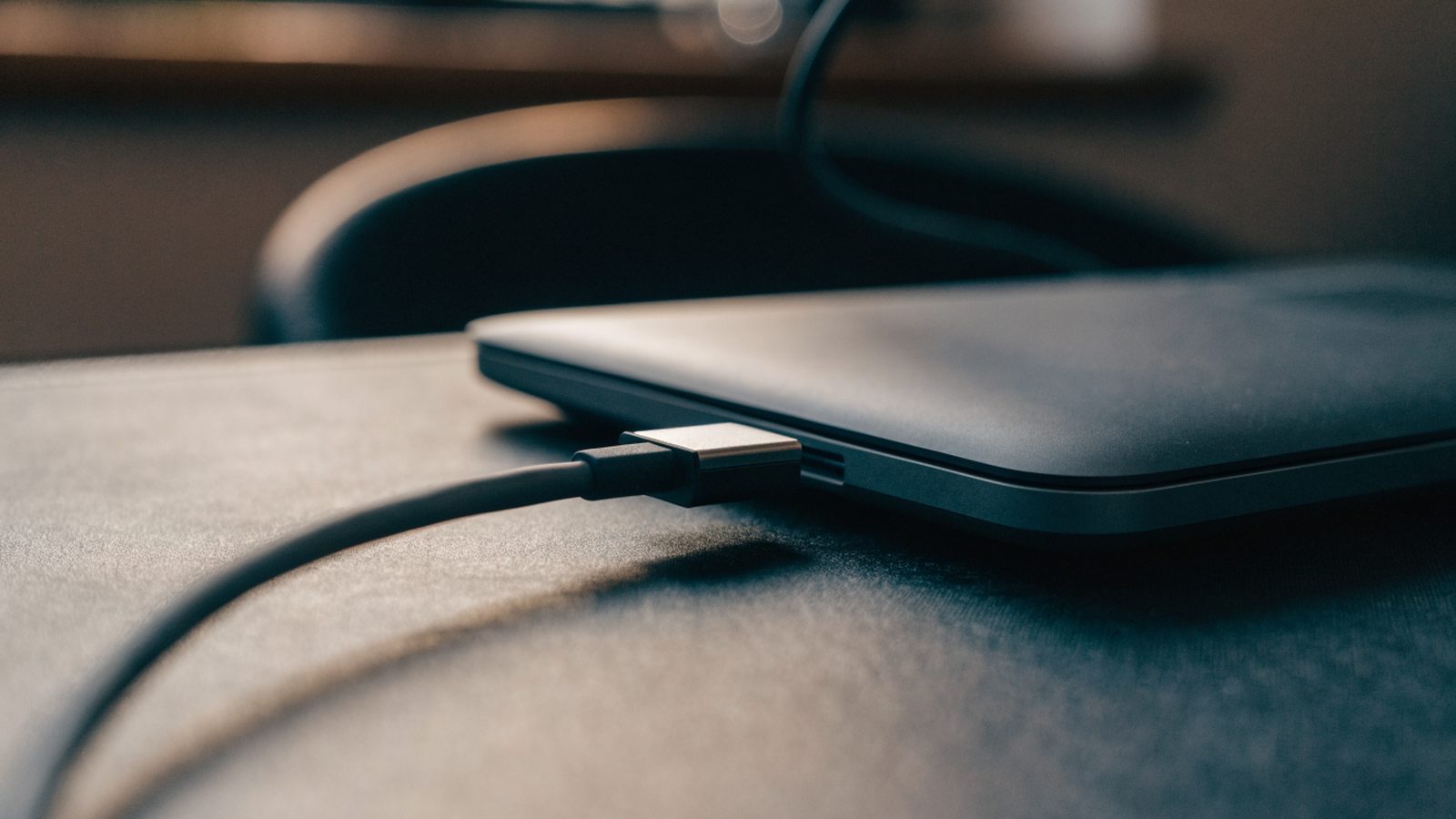 Close-up of a USB-C charger cable plugged into the side port of a graphite black laptop on a dark desk mat