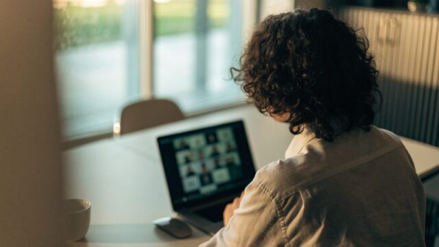 A person seen from behind on a video call on a graphite black laptop on an office desk