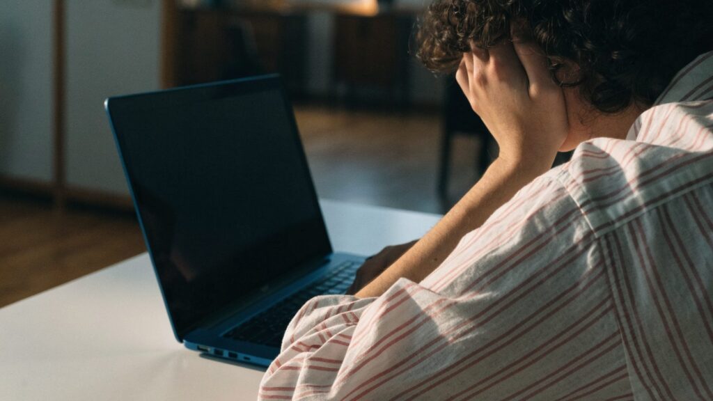 A person resting their forehead on one hand in front of a dark blue laptop on a white desk
