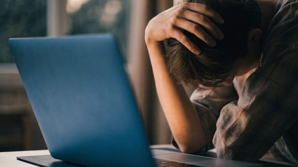 A person resting their forehead on one hand in front of a dark blue laptop on a dark desk mat