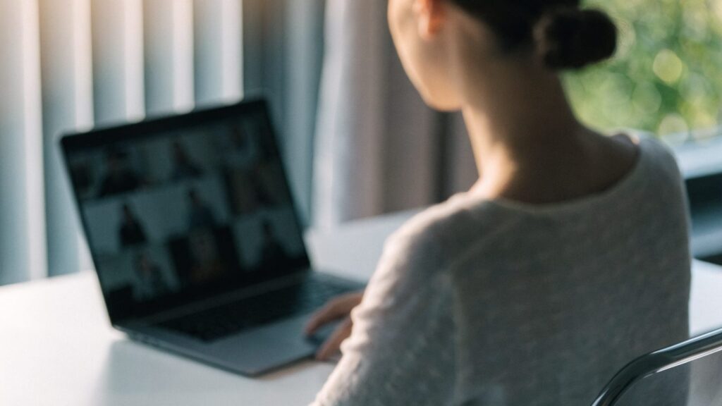 A person seen from behind on a video call on a matte grey laptop on a white desk