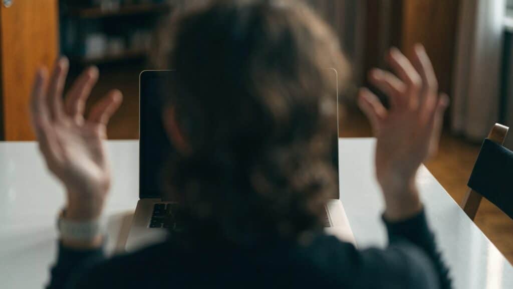A person seen from behind with hands raised at a silver laptop with a dark blank screen on a white desk