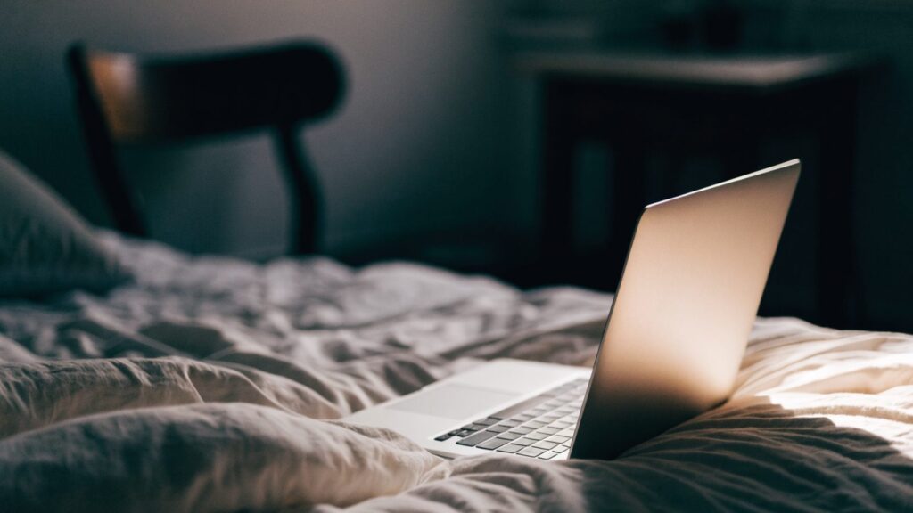 A silver laptop glowing softly on a bed in a dark room