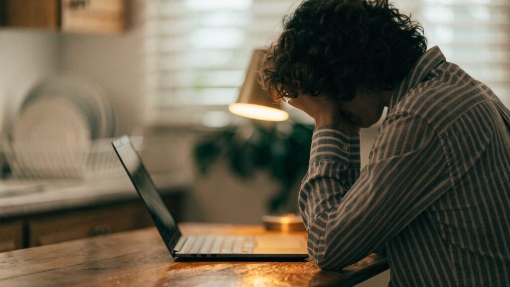 A person resting their forehead on one hand in front of a graphite black laptop on a kitchen table