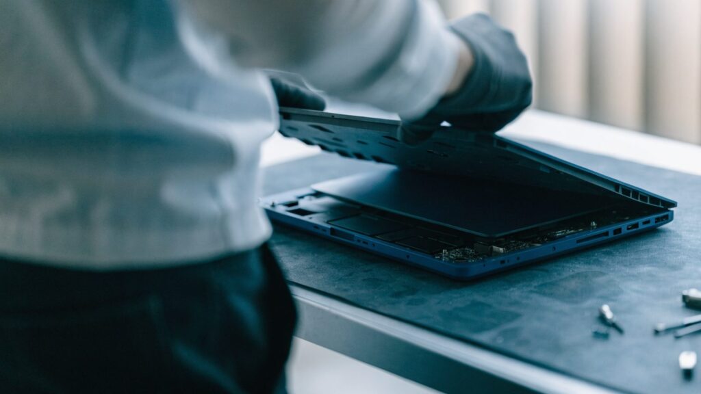 A technician lifting the bottom cover off a dark blue laptop on an anti-static mat
