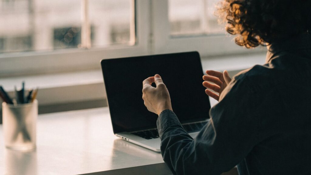 A person seen from behind with hands raised at a silver laptop with a dark blank screen on a white desk