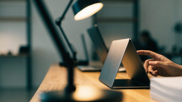 A hand touching the screen of a convertible matte grey laptop folded in tent mode on an office desk
