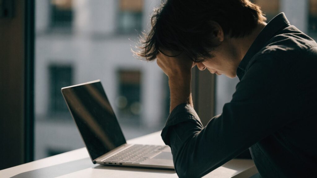 A person resting their forehead on one hand in front of a matte grey laptop on a white desk