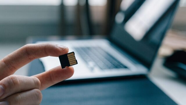 An SD card held between two fingers in front of a silver laptop on a dark desk mat