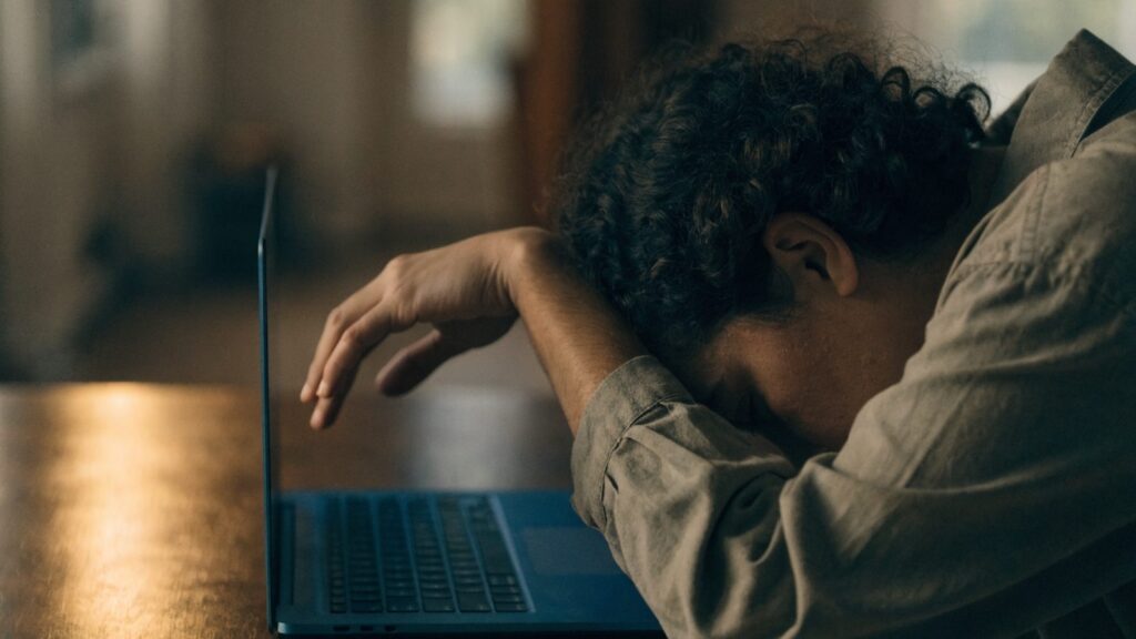 A person resting their forehead on one hand in front of a dark blue laptop on a wooden desk