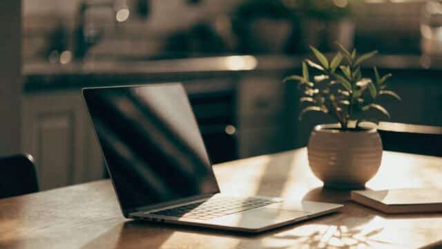 A silver laptop open on a kitchen table with a small plant and notebook beside it