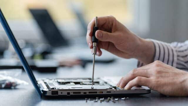 A technician's hands holding a small screwdriver over an opened dark blue laptop with internal components visi