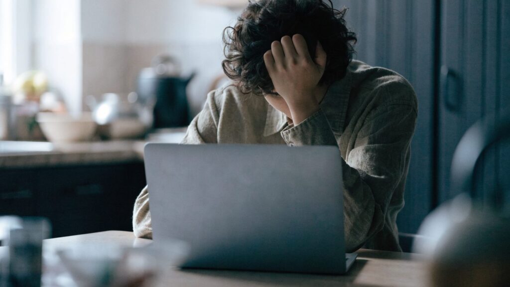 A person resting their forehead on one hand in front of a matte grey laptop on a kitchen table