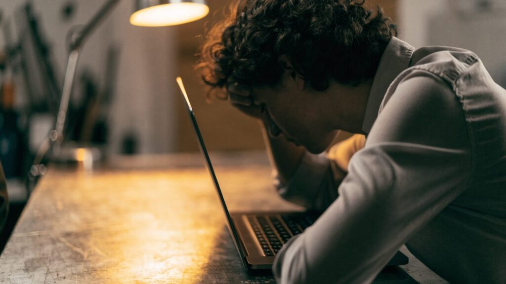 A person resting their forehead on one hand in front of a graphite black laptop on a workbench