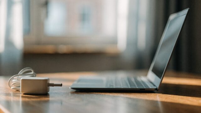 A matte grey laptop on a wooden desk next to a compact white power adapter with its cable neatly coiled