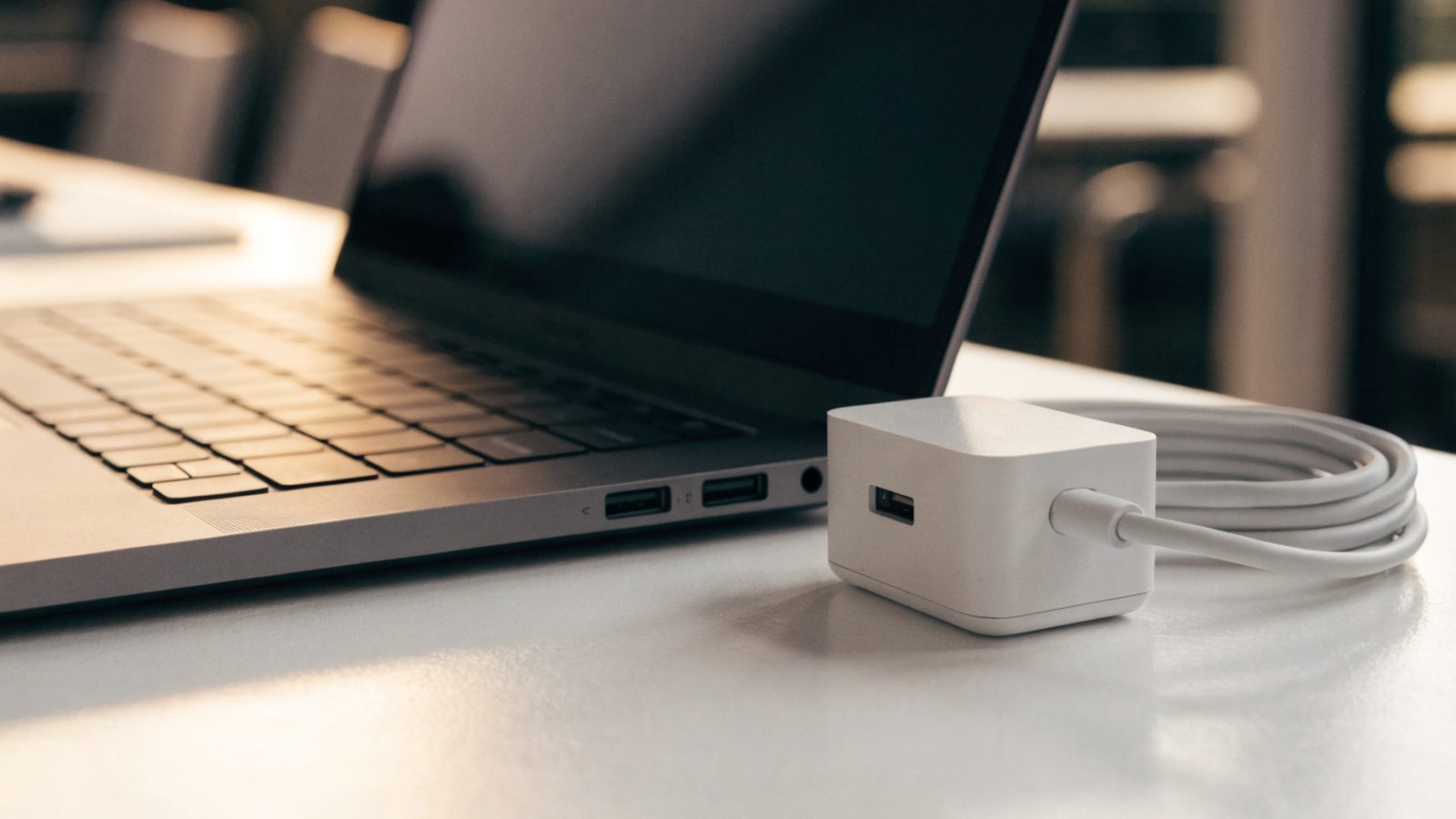 A matte grey laptop on a white desk next to a compact white power adapter with its cable neatly coiled