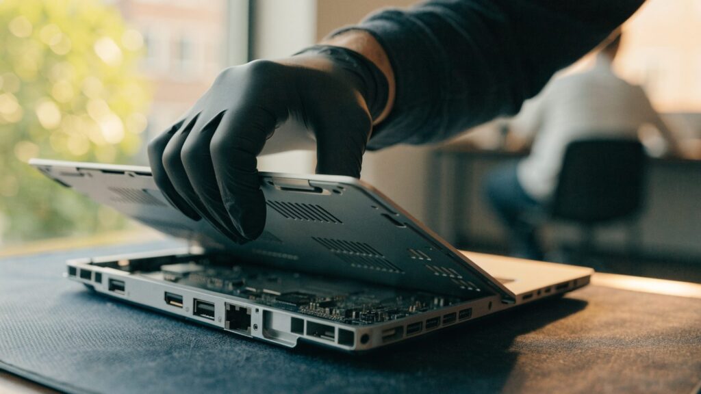 A technician lifting the bottom cover off a silver laptop on an anti-static mat
