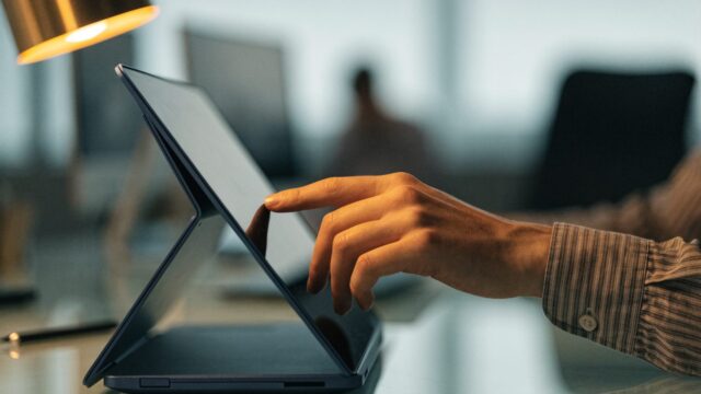 A hand touching the screen of a convertible dark blue laptop folded in tent mode on an office desk
