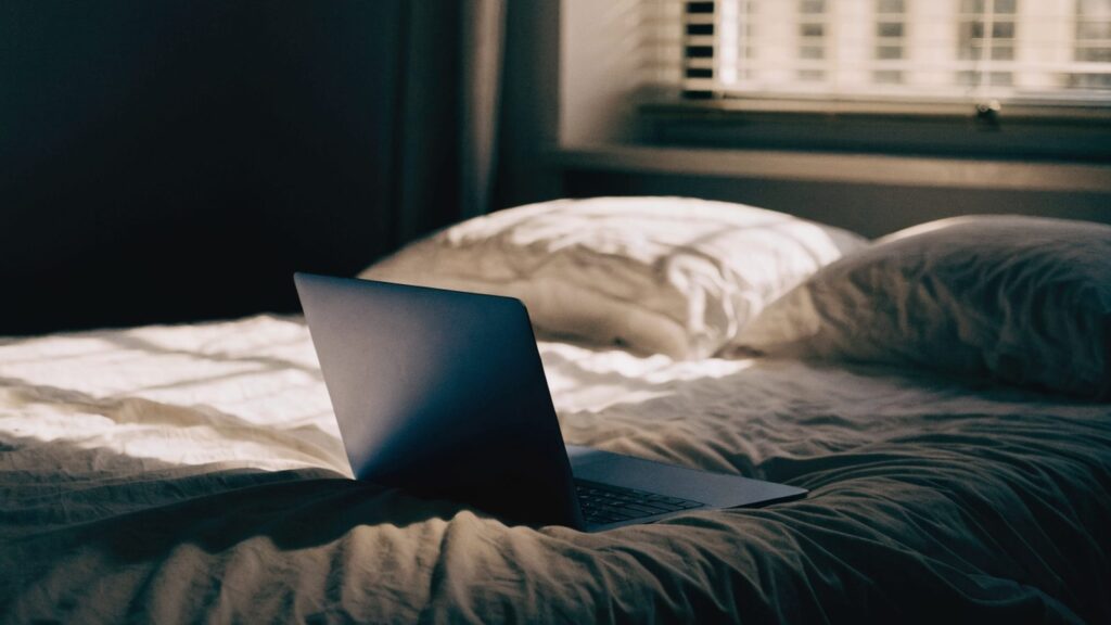 A dark blue laptop glowing softly on a bed in a dark room