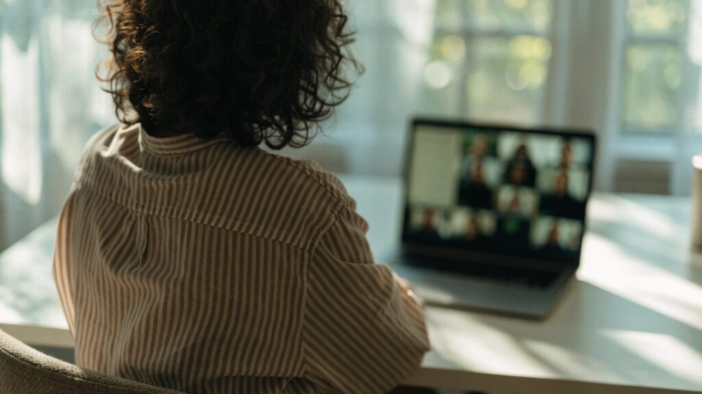 A person seen from behind on a video call on a matte grey laptop on a white desk