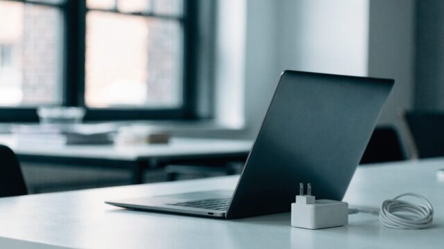 A graphite black laptop on an office desk next to a compact white power adapter with its cable neatly coiled