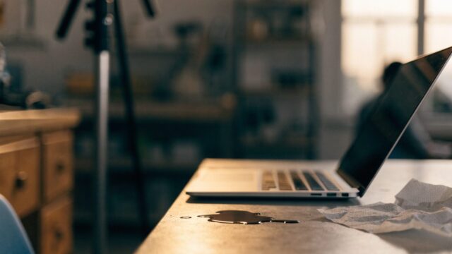 A small puddle of spilled coffee next to a silver laptop on a workbench