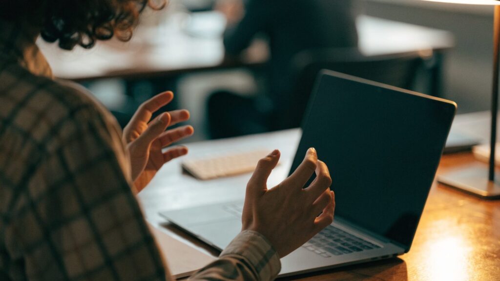 A person seen from behind with hands raised at a matte grey laptop with a dark blank screen on an office desk