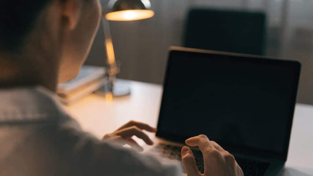 A person seen from behind with hands raised at a matte grey laptop with a dark blank screen on a white desk