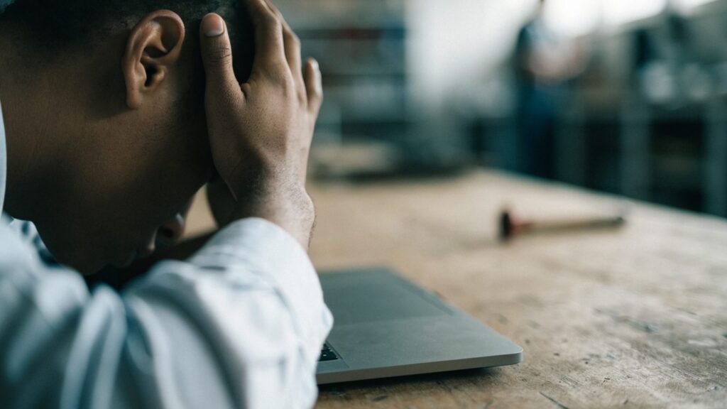 A person resting their forehead on one hand in front of a matte grey laptop on a workbench