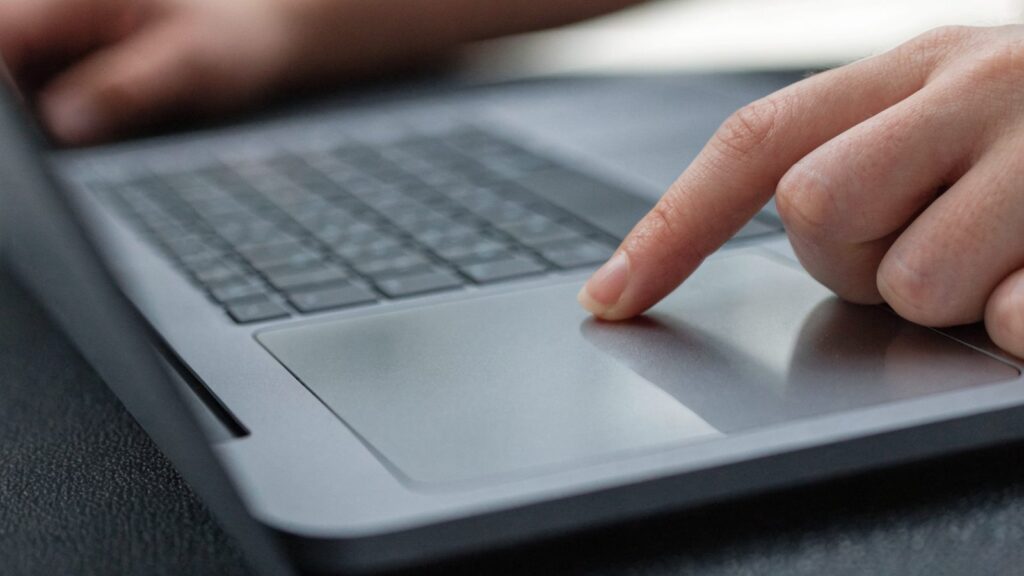 A finger gliding on the touchpad of a matte grey laptop on a dark desk mat