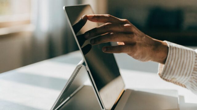 A hand touching the screen of a convertible silver laptop folded in tent mode on a white desk