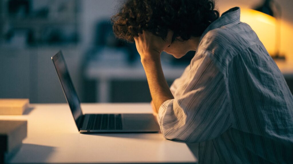 A person resting their forehead on one hand in front of a matte grey laptop on a white desk
