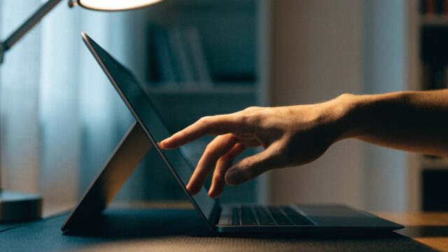 A hand touching the screen of a convertible graphite black laptop folded in tent mode on a dark desk mat