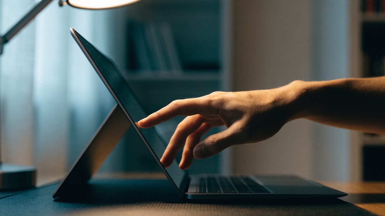 A hand touching the screen of a convertible graphite black laptop folded in tent mode on a dark desk mat