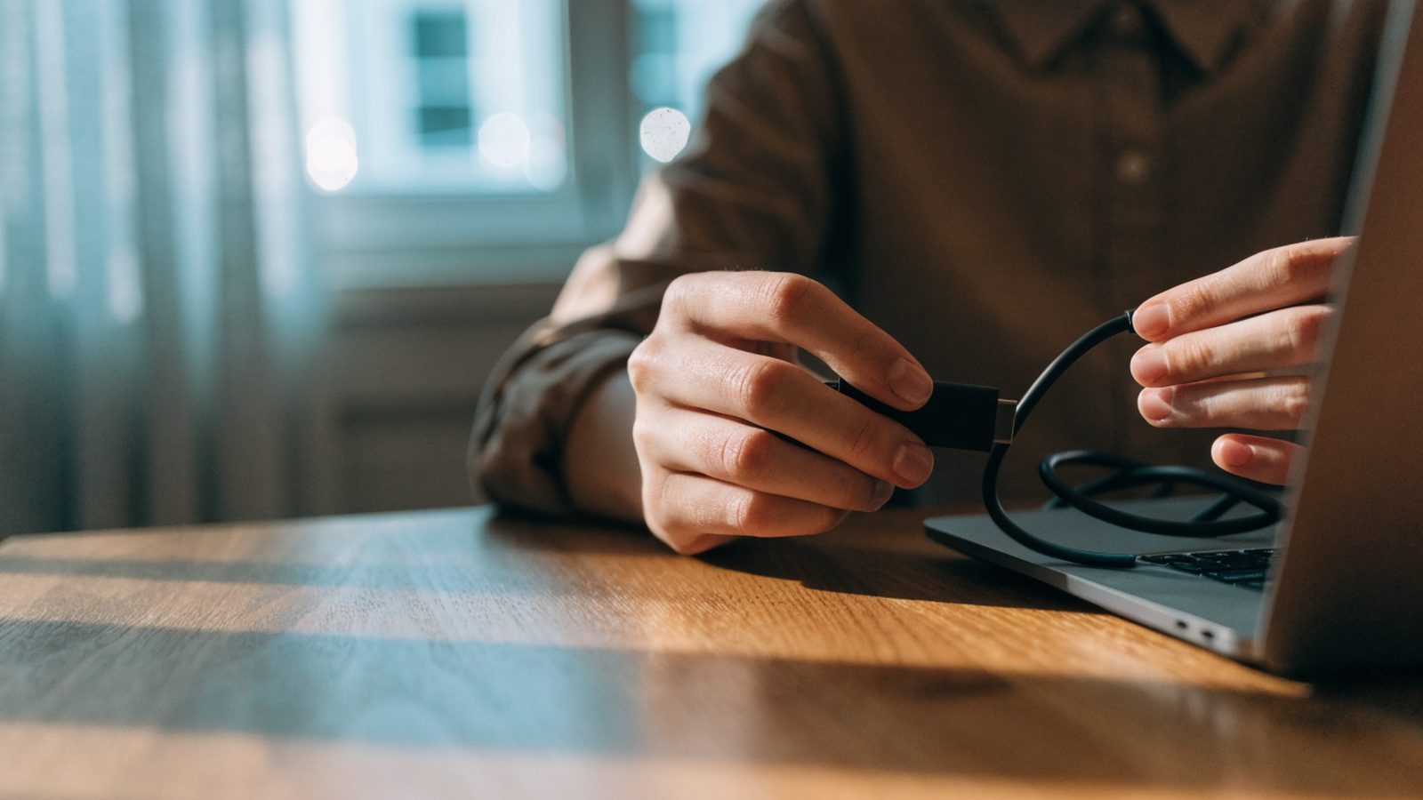 Hands unplugging a charging cable from a slim matte grey laptop on a wooden desk