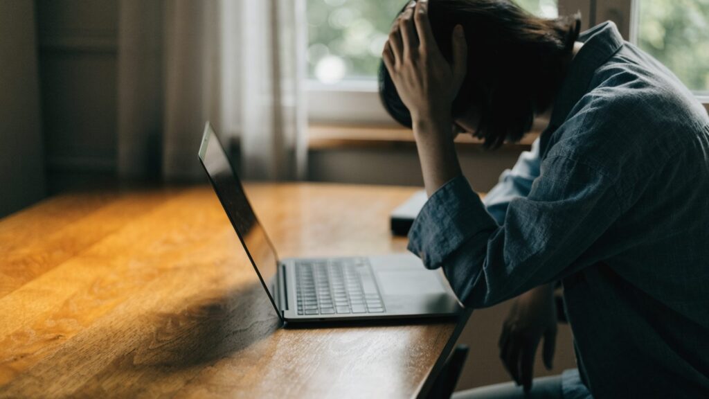 A person resting their forehead on one hand in front of a graphite black laptop on a wooden desk