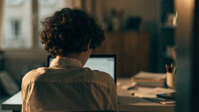 A teenager seen from behind doing homework on a matte grey laptop at a desk