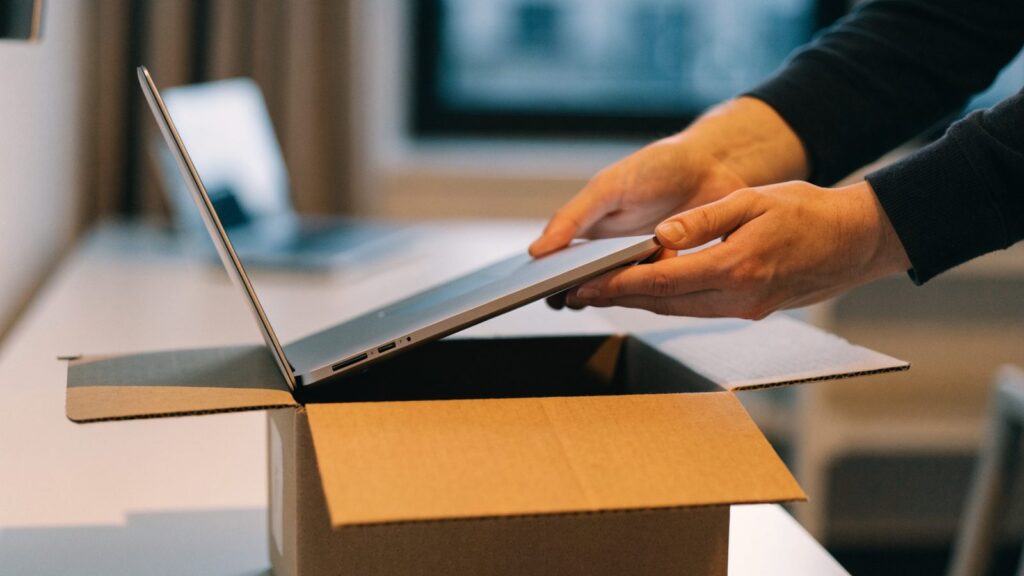 Hands lifting a brand-new silver laptop out of its plain cardboard box on a white desk