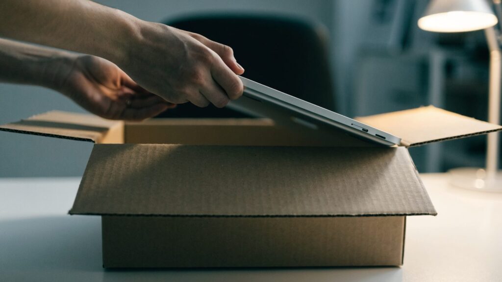 Hands lifting a brand-new silver laptop out of its plain cardboard box on a white desk