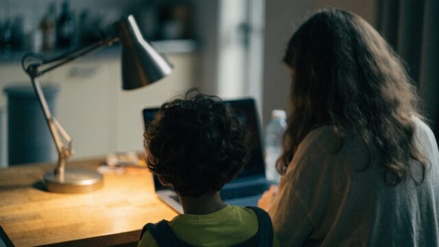 A parent and child seen from behind using a dark blue laptop together at a kitchen table