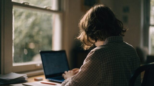 A teenager seen from behind doing homework on a dark blue laptop at a desk