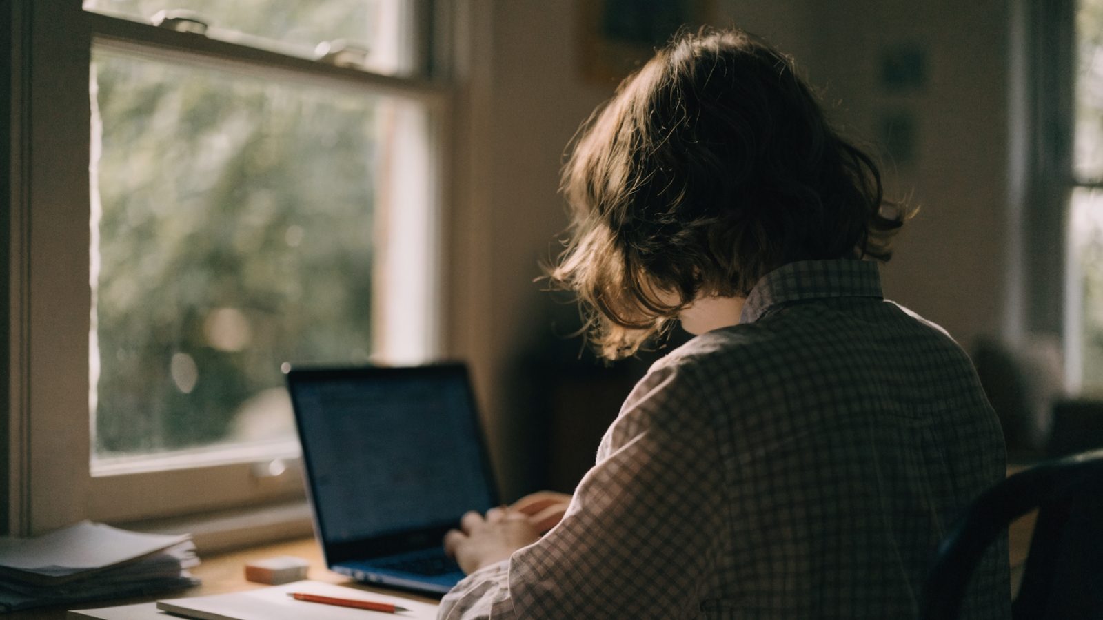 A teenager seen from behind doing homework on a dark blue laptop at a desk