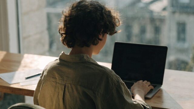 A teenager seen from behind doing homework on a matte grey laptop at a desk